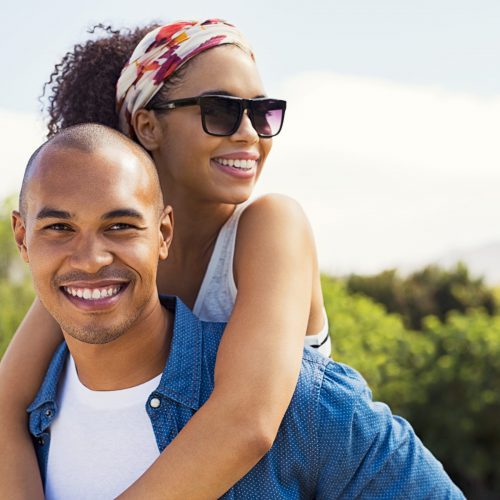 Portrait of young black man holding beautiful woman on back and looking at camera. African american woman with sunglasses enjoying piggyback on boyfriend at park and looking away in a summer holiday.