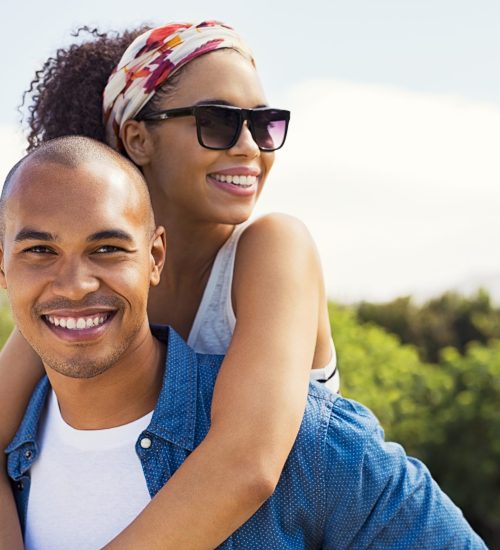 Portrait of young black man holding beautiful woman on back and looking at camera. African american woman with sunglasses enjoying piggyback on boyfriend at park and looking away in a summer holiday.